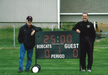 Coach Duke (right) stands next to the scoreboard with his dad, who used to keep score.