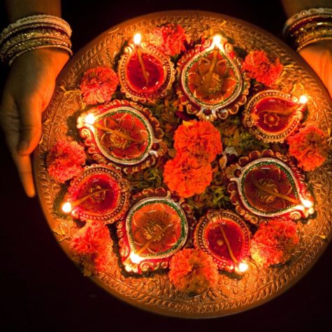 A plate of lit diyas arranged in a floral pattern with marigolds being held by a woman adorned with gold bangles.
