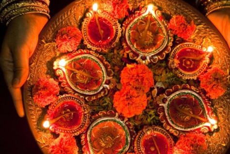 A plate of lit diyas arranged in a floral pattern with marigolds being held by a woman adorned with gold bangles.
